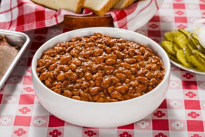 Bowl of saucy baked beans on a red checkered picnic tablecloth with pickles on the side