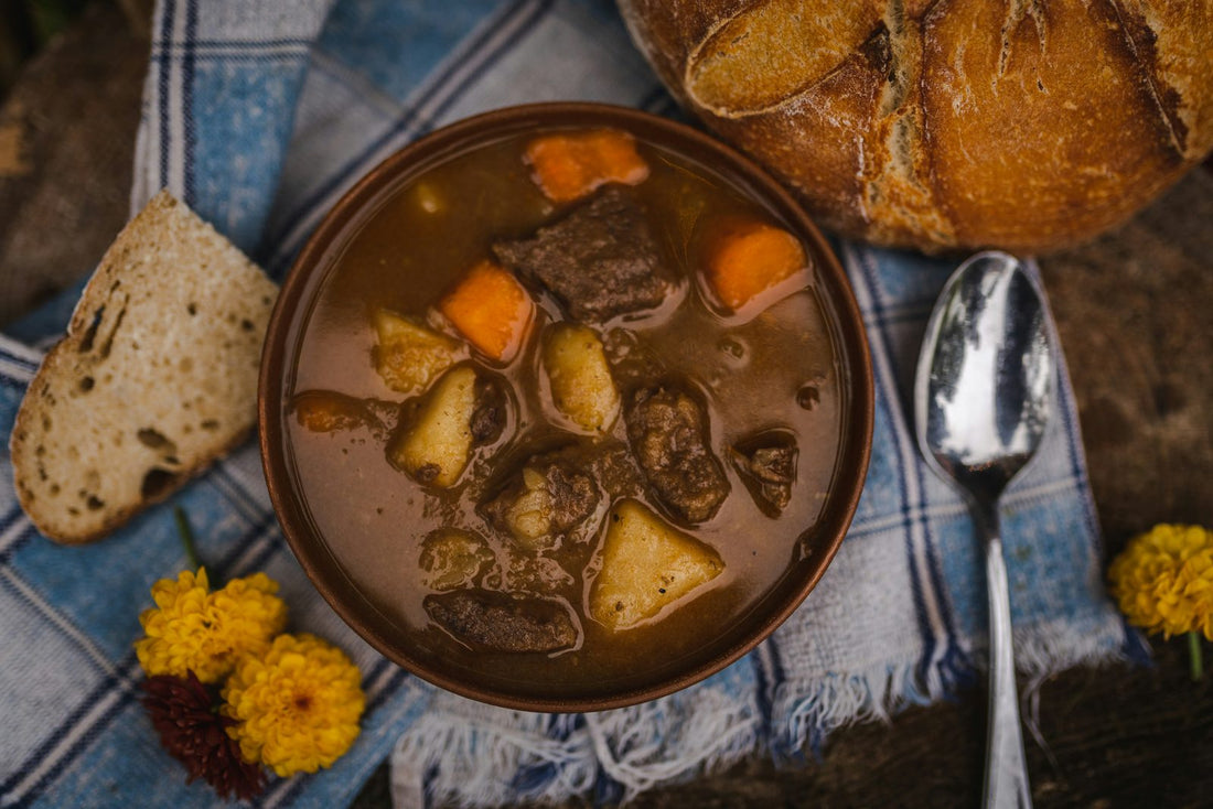 Bowl of beef stew with carrots and potatoes beside crusty bread on a blue checked cloth