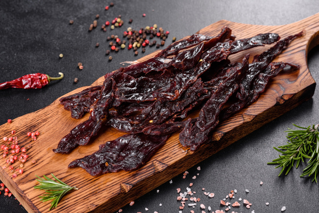 Dark beef jerky on an olive-wood board with peppercorns