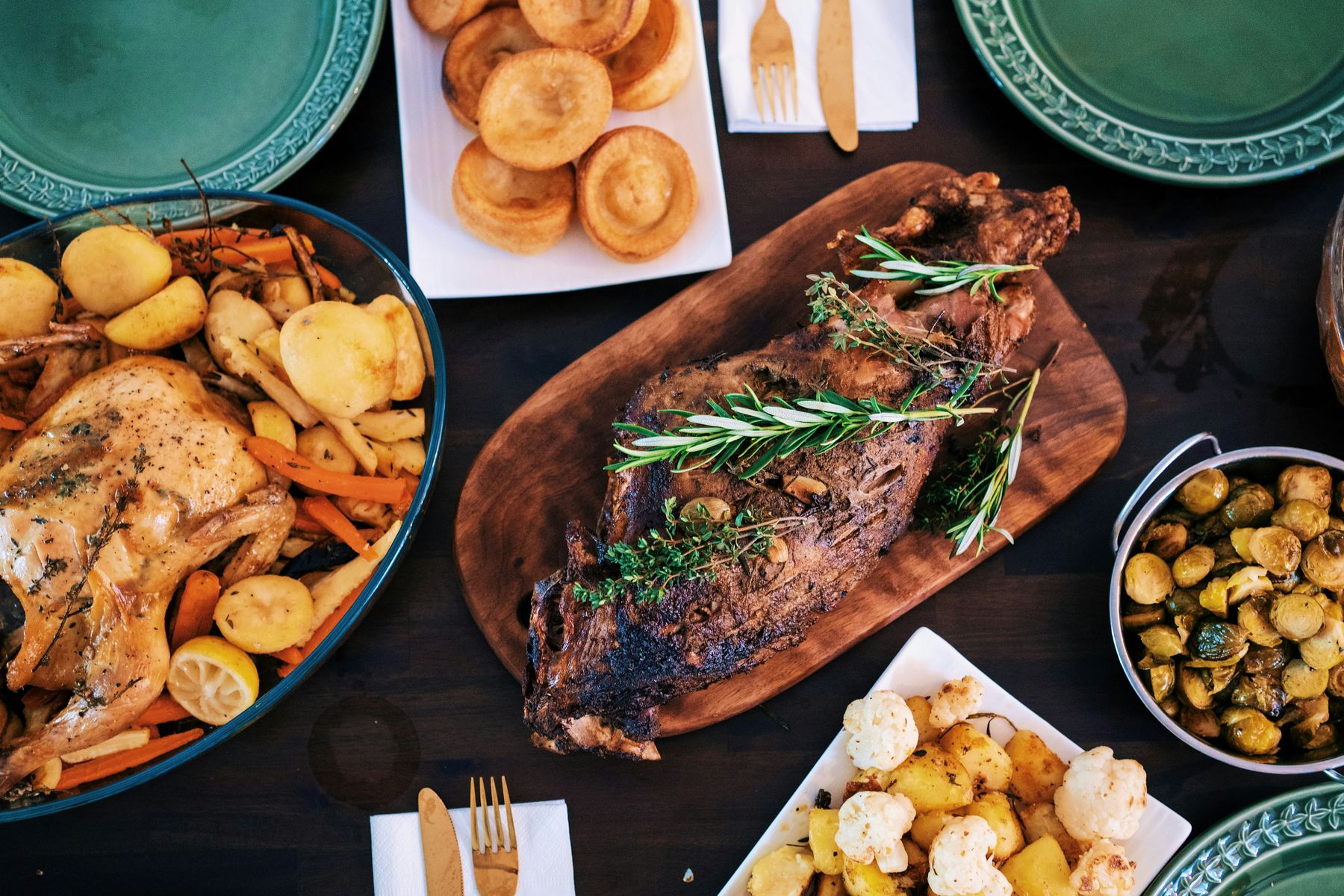 Roast dinner spread with a herb-topped roast on a wooden board, roast chicken with potatoes, and sides