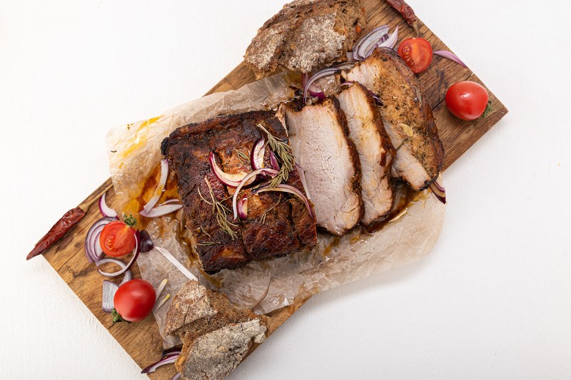 Sliced roast pork on a wooden board with parchment, red onion, and cherry tomatoes