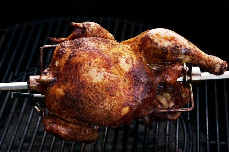 Whole seasoned chicken turning on a rotisserie spit over the grill