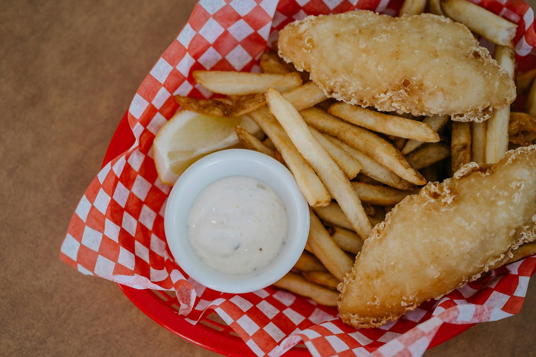 Crisp fried fish fillets with fries, lemon and tartar sauce in a checkered paper basket